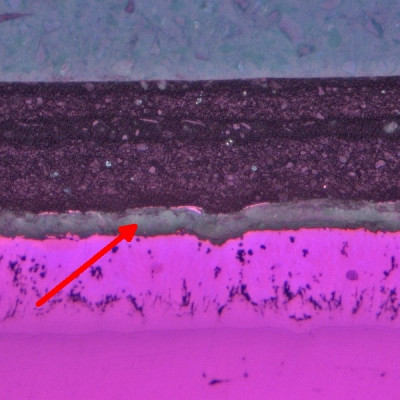 Metallographischer Schliff: zwischen der Feuerverzinkung und der Beschichtung ist Weißrost erkennbar (Pfeil); Vergr.: 100:1